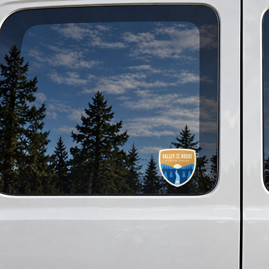 Valley of the Rogue sticker on the rear window of a van, reflecting conifers and blue sky